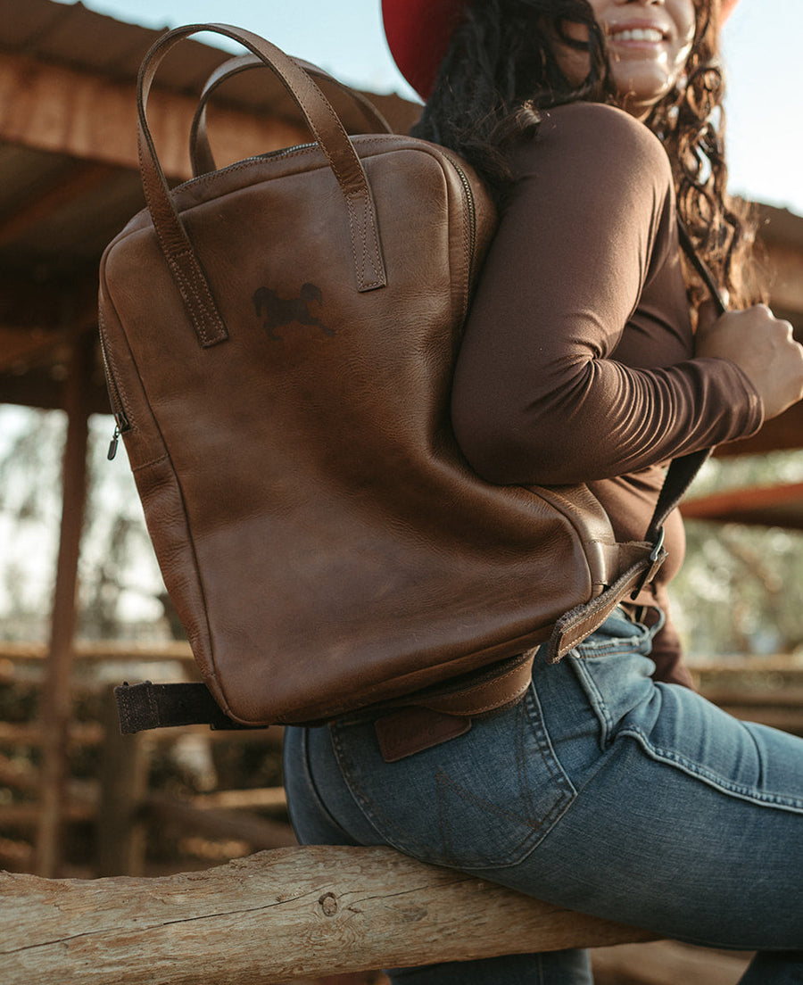 The Margaux leather backpack worn by a woman sitting on a fence rail