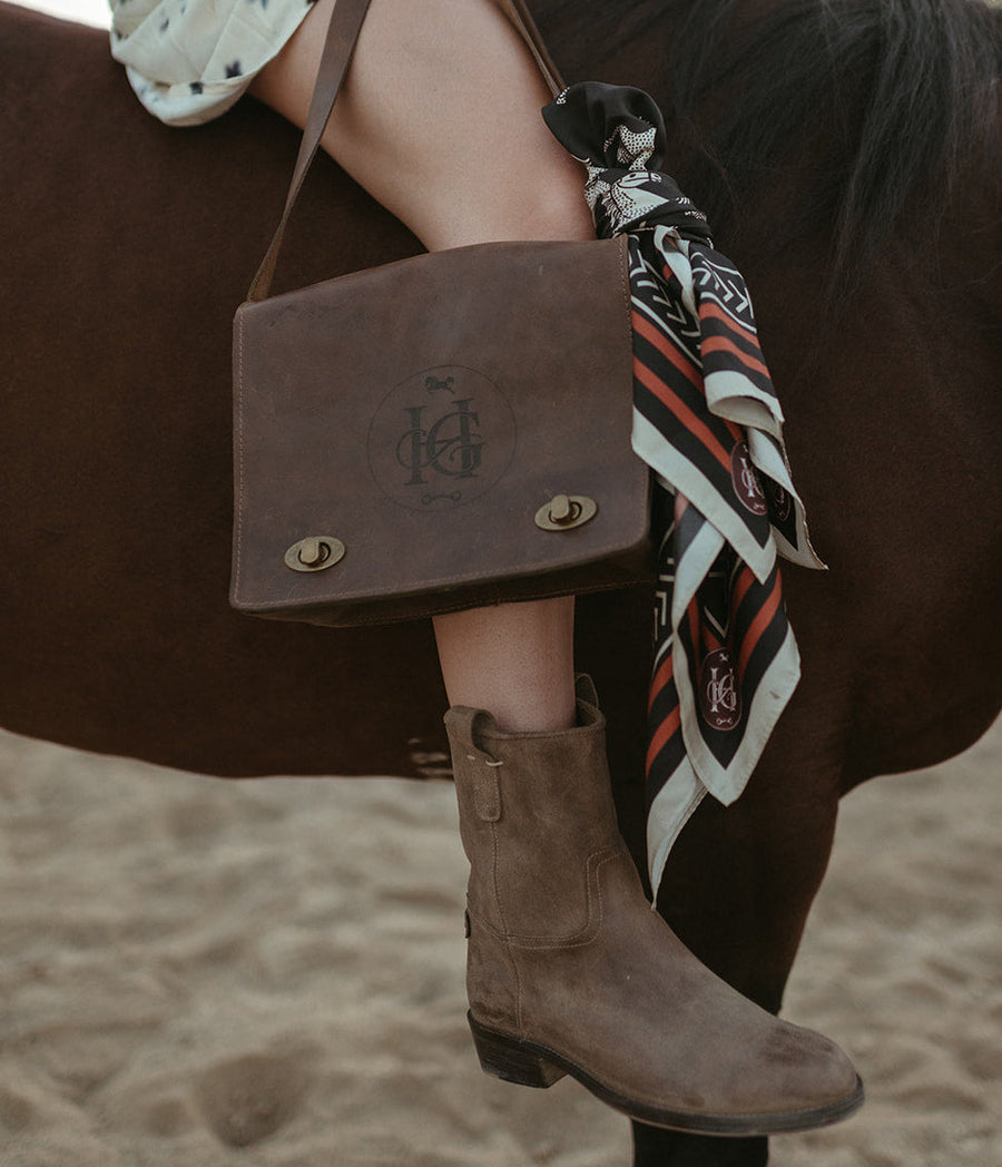 The Eleanora saddle bag worn crossbody by a rider, with The Claudia boot and the Horse Girl scarf