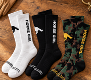 The Iconic Horse Girl Boot Sock in white, black and camouflage laid out on wood