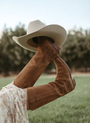 The Tahlia tall suede boots raised in the air with a cowboy hat on the toe
